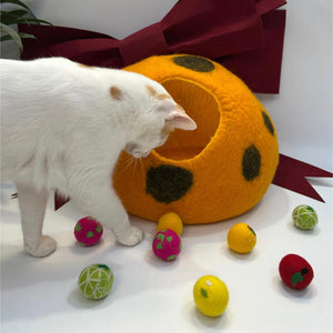 Vibrant wool felt fruit balls scattered near a large orange cat cave bed. The playful cat is exploring the handcrafted toys, making a stylish pet gift set.