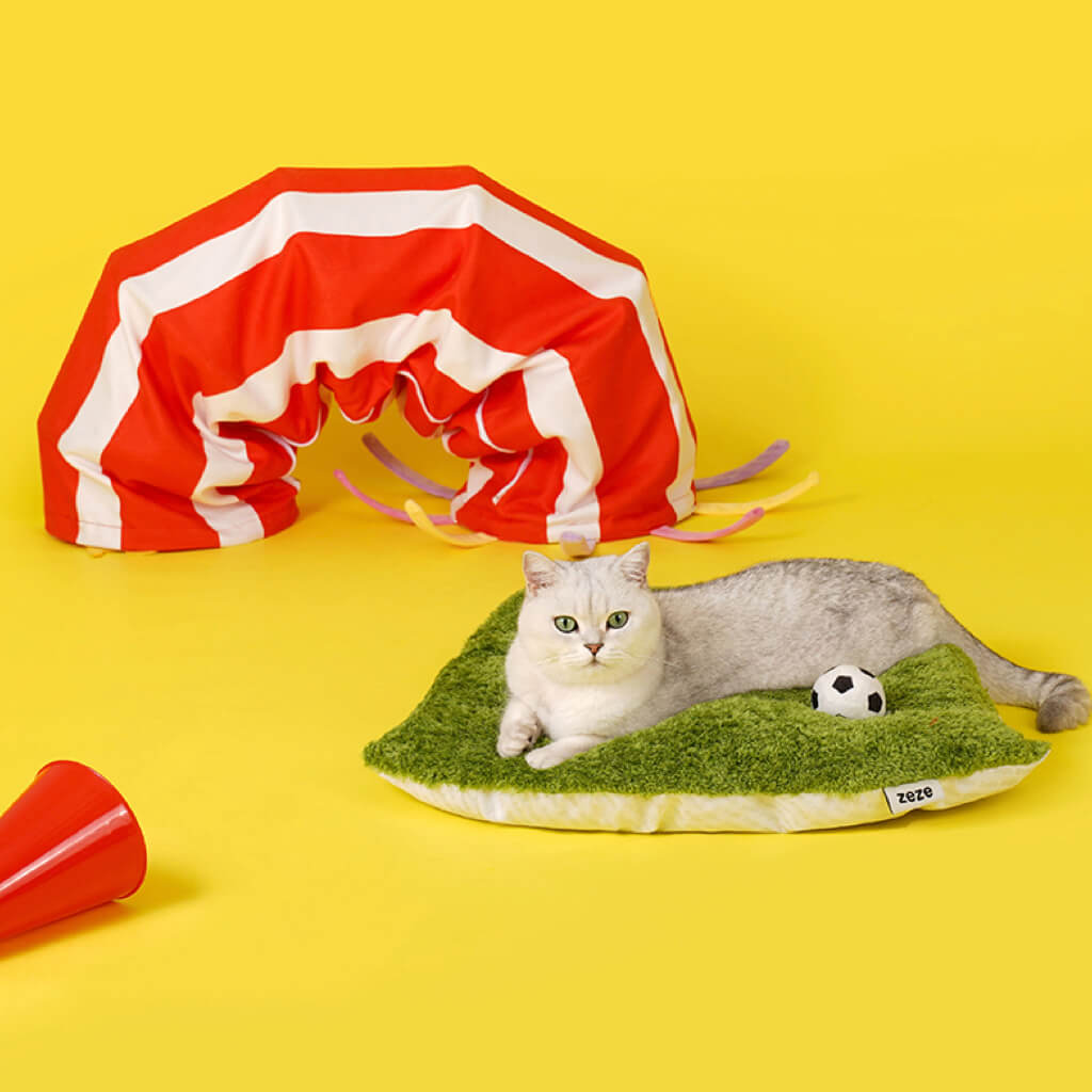 Cat lounging on the plush grass-textured bed of the ZEZE Football Field Cat Tunnel Toy, with the tunnel in the background.