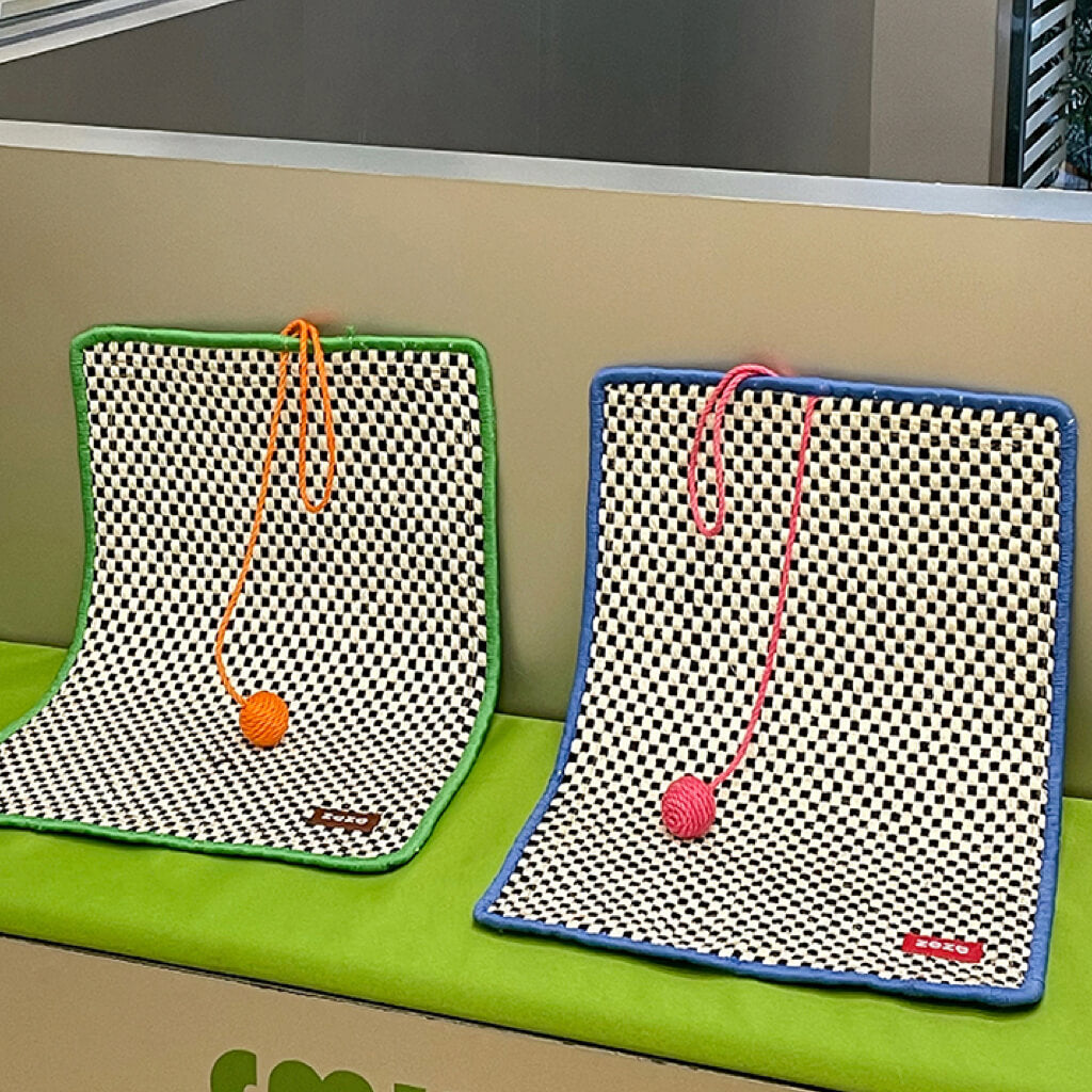 Two retro-style cat sisal mats in green and blue, with contrasting pom-poms, displayed on a green surface.