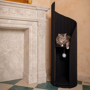 Cat lounging comfortably on the elevated perch of the Time Travel Cat Tower - Black next to a marble fireplace.