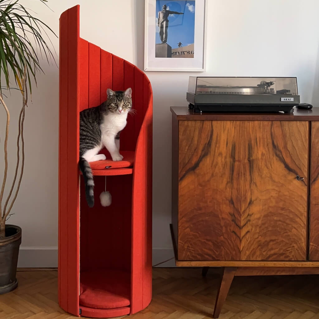 A tabby cat sitting on the top platform of the red Time Travel Cat Tower in a retro-style room. The tower stands next to a mid-century modern cabinet with wood grain detailing, and framed photographs on the wall create a chic yet cozy vibe for both the cat and its owner.