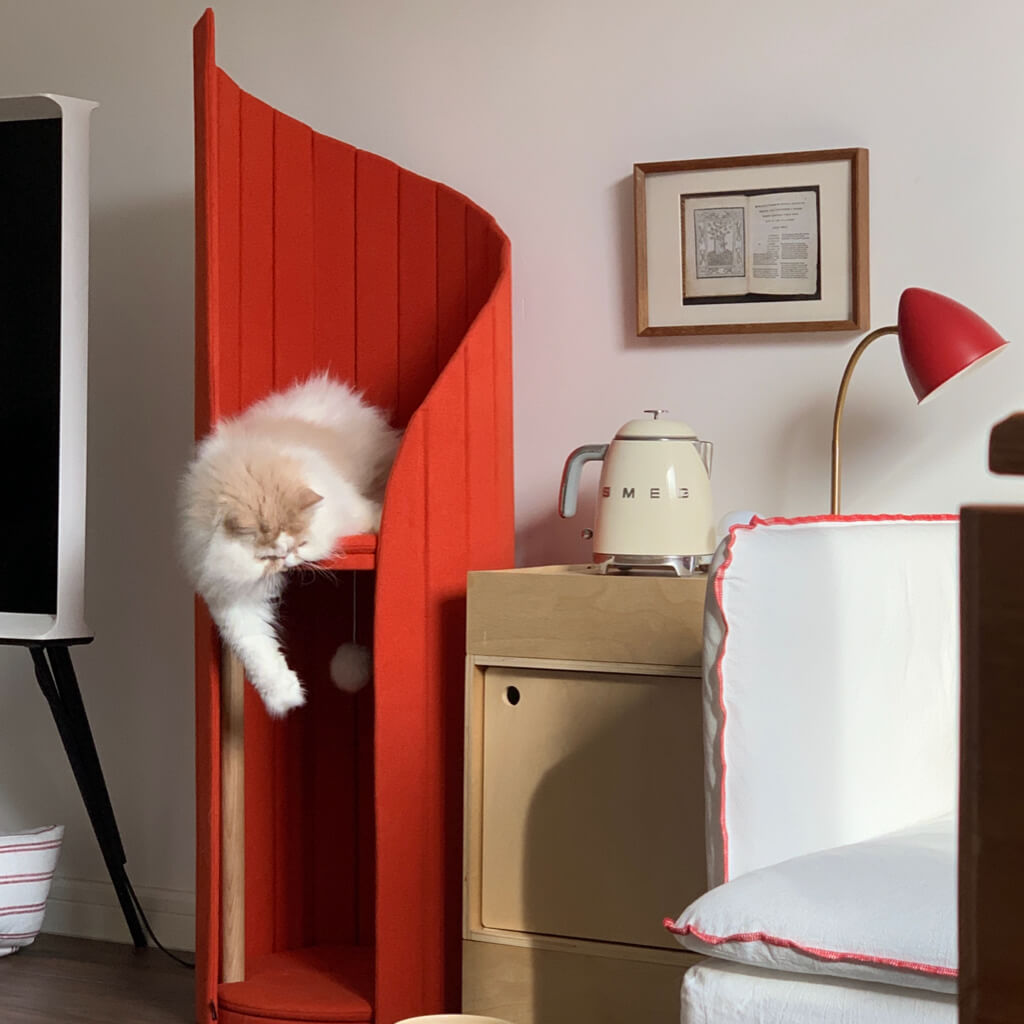 A fluffy white cat is comfortably sprawled across the top of the red Time Travel Cat Tower, with one paw hanging lazily over the edge. The tower is placed next to a light wooden nightstand and small minimalist pet bowls, fitting seamlessly into the cozy bedroom setting.